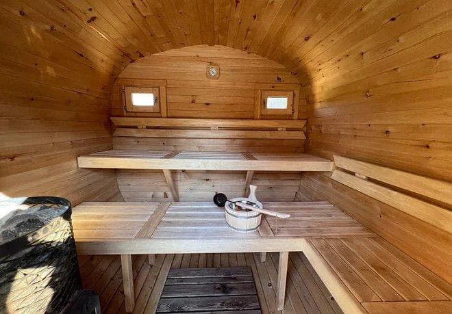 Innenansicht einer Fasssauna aus Holz mit Ofen, Aufgusseimer und zwei Sitzebenen für Wellness und Entspannung.